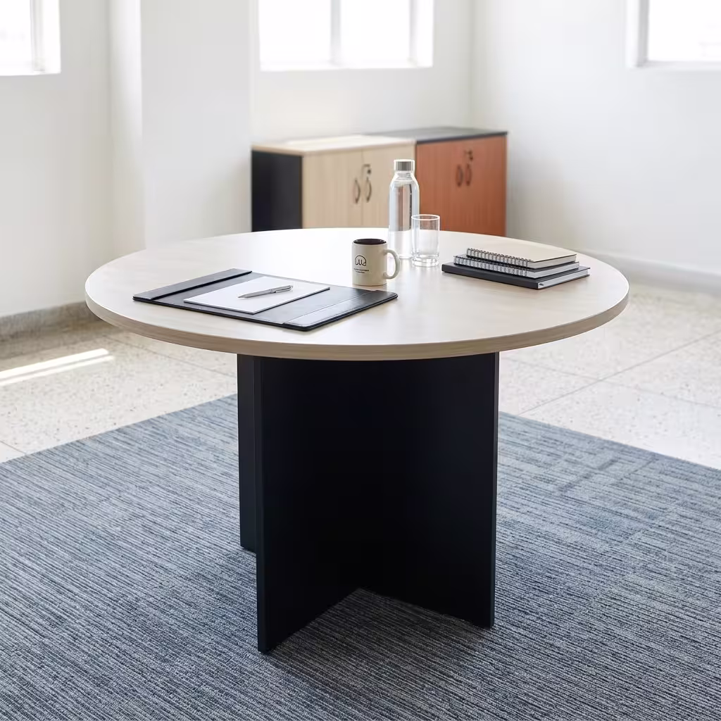 Premium 1.4M Round Meeting Table in a light oak wood finish with a black metal cross-pedestal base, paired with modern grey mesh chairs in a sunlit meeting room.