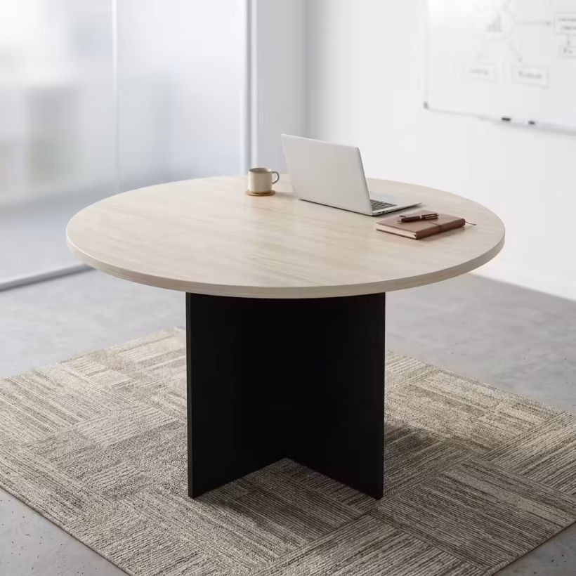 Premium 1.4M Round Meeting Table in a light oak wood finish with a black metal cross-pedestal base, paired with modern grey mesh chairs in a sunlit meeting room.