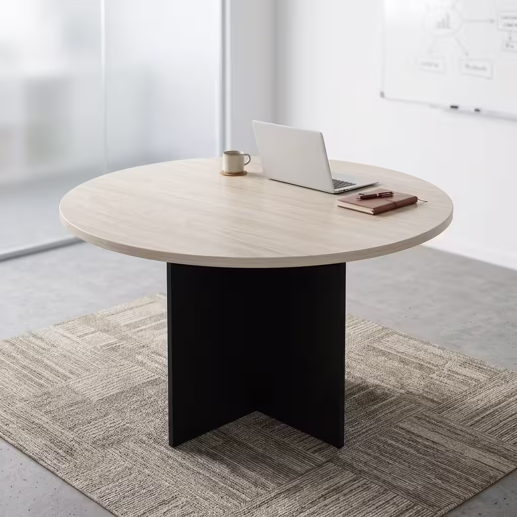 Premium 1.4M Round Meeting Table in a light oak wood finish with a black metal cross-pedestal base, paired with modern grey mesh chairs in a sunlit meeting room.
