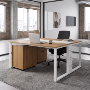 Premium 2-Way Modular Office Workstation featuring two back-to-back light woodgrain desktops, a central white privacy strip, white steel loop base legs, an integrated woodgrain drawer pedestal with a key lock, displaying a keyboard, a laptop, and a desk lamp on a geometric grey carpet.