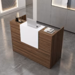 Premium 1.2M Wooden Reception Counter Office Desk featuring a rich walnut woodgrain panel facade, a center-set cascading waterfall-edge white transaction counter supporting a silver laptop, a pen holder set, and stacked books, resting on a polished grey concrete floor.