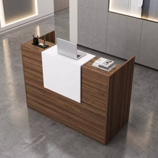Premium 1.2M Wooden Reception Counter Office Desk featuring a rich walnut woodgrain panel facade, a center-set cascading waterfall-edge white transaction counter supporting a silver laptop, a pen holder set, and stacked books, resting on a polished grey concrete floor.