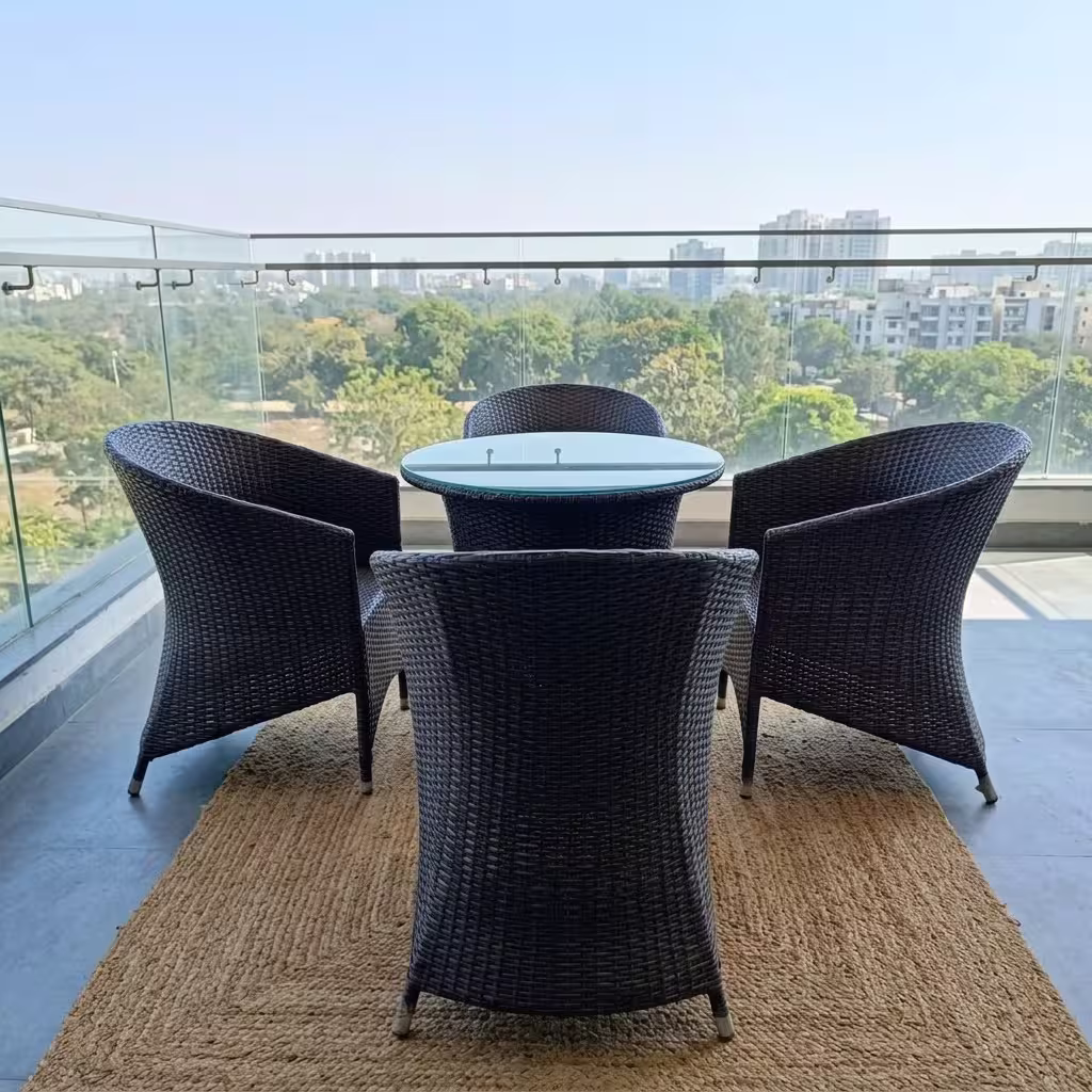 Luxury 4-Seater Rattan Outdoor furniture set featuring four dark hand-woven wicker armchairs and a circular glass-top table on a woven mat on a scenic balcony.