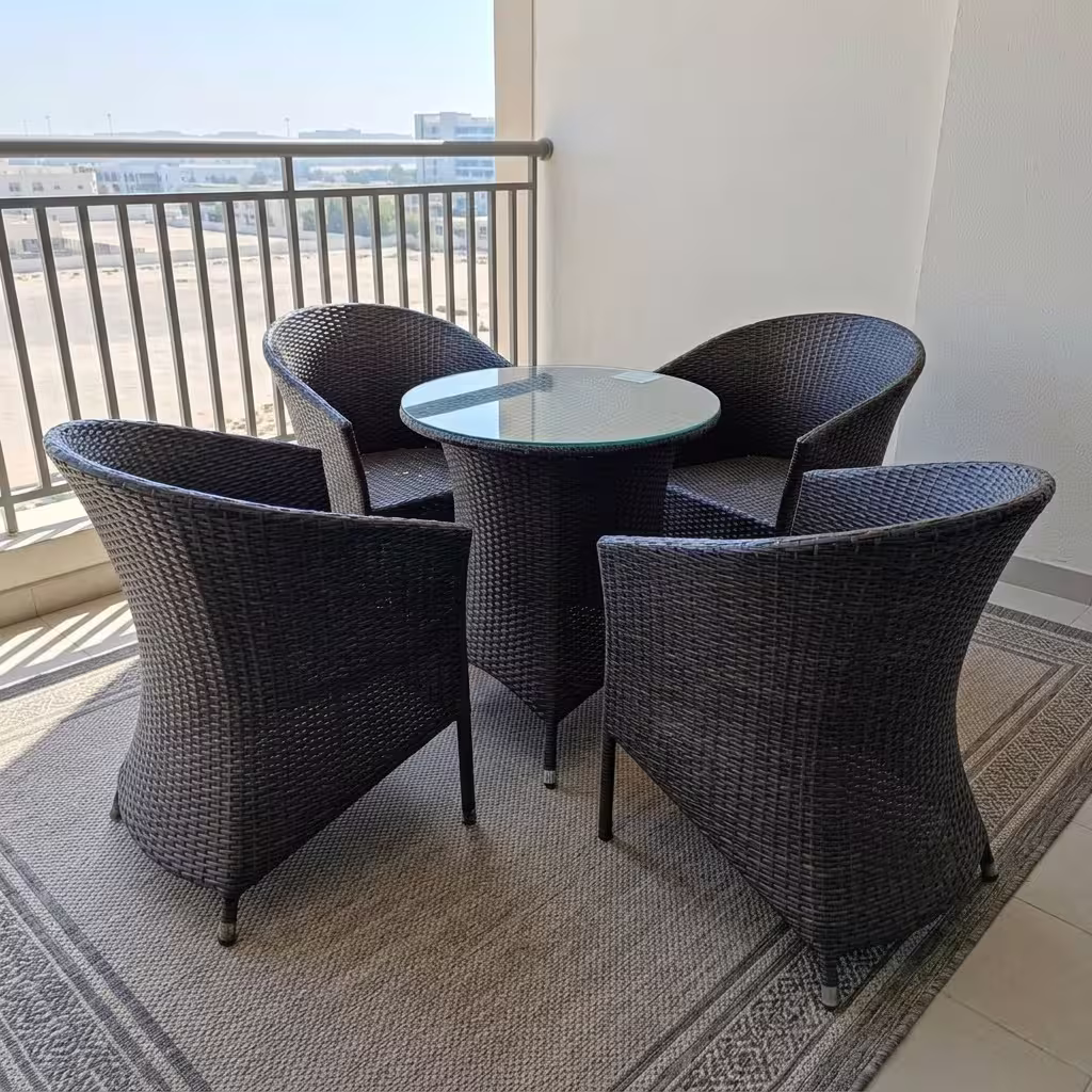 Luxury 4-Seater Rattan Outdoor furniture set featuring four dark hand-woven wicker armchairs and a circular glass-top table on a woven mat on a scenic balcony.