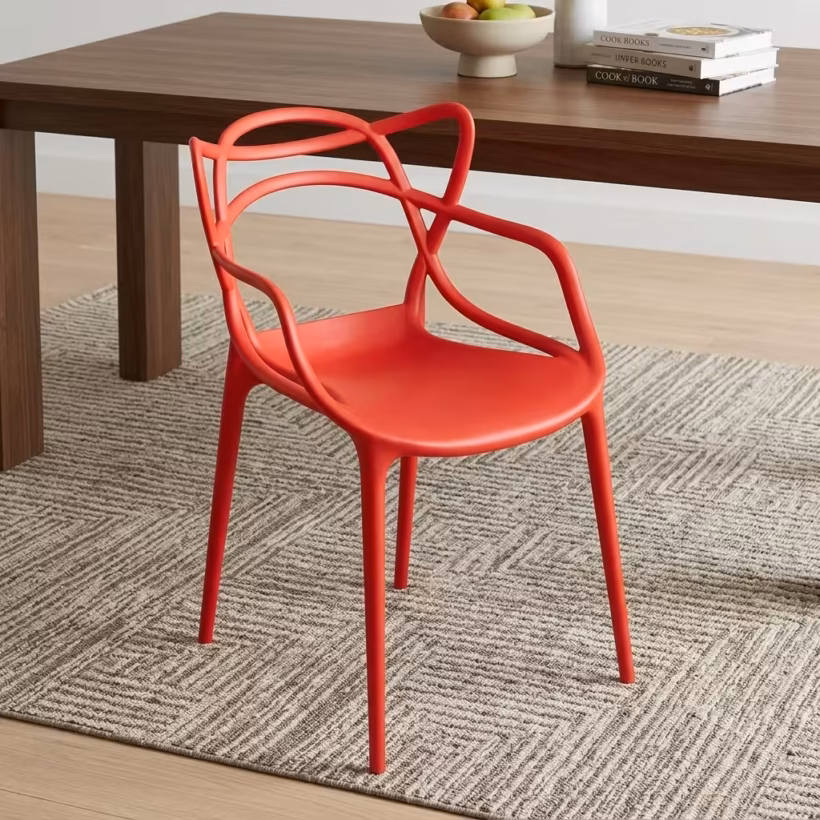 Premium Modern Plastic Outdoor Dining Chair in a vibrant coral-red matte finish, showcasing an intricate interlocking loop open-back silhouette, integrated sweeping armrests, and matching tapered legs, resting on a textured geometric gray area rug next to a walnut wood dining table.