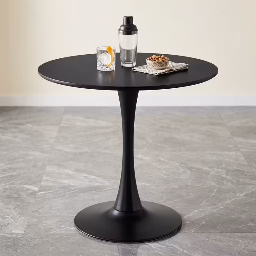Premium Modern Round Cocktail Table featuring a smooth circular matte black tabletop, supported by a matching matte black trumpet pedestal base, displaying a glass and cocktail shaker on a grey marble tile floor.