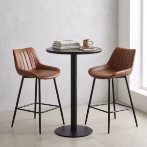 Premium Home Bar Cocktail Table Set featuring a round matte black pedestal table holding a stack of books and a mug, flanked by two matching biscuit-stitched brown faux leather bar stools with black steel frames.