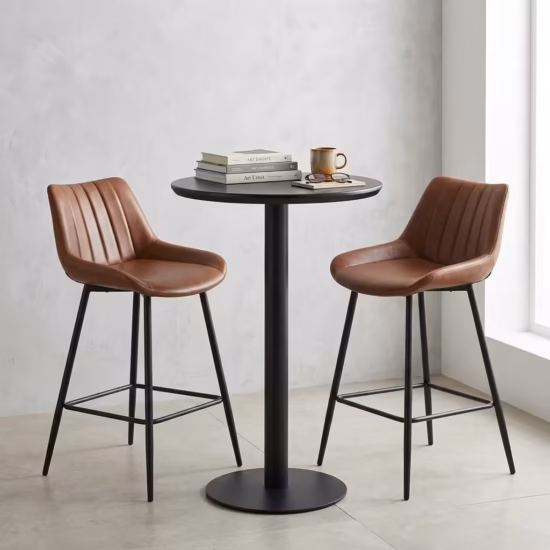 Premium Home Bar Cocktail Table Set featuring a round matte black pedestal table holding a stack of books and a mug, flanked by two matching biscuit-stitched brown faux leather bar stools with black steel frames.
