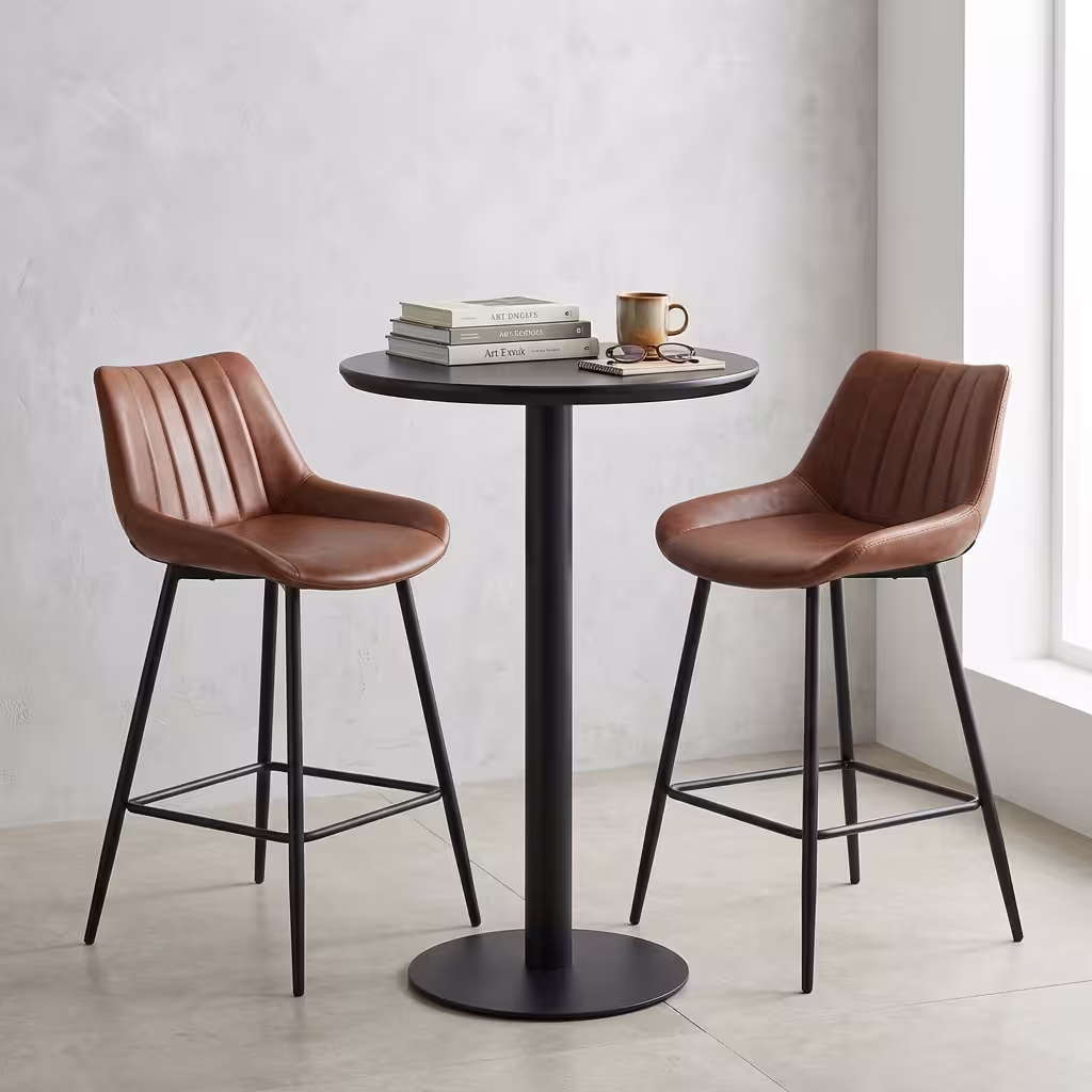 Premium Home Bar Cocktail Table Set featuring a round matte black pedestal table holding a stack of books and a mug, flanked by two matching biscuit-stitched brown faux leather bar stools with black steel frames.