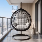 Premium 1-Seater Modern Rattan Hammock Swing featuring a black hand-woven teardrop wicker basket, a heavy-duty dark gray steel hanging stand, and plush black-and-white plaid cushions, placed on a high-rise balcony overlooking a city skyline.