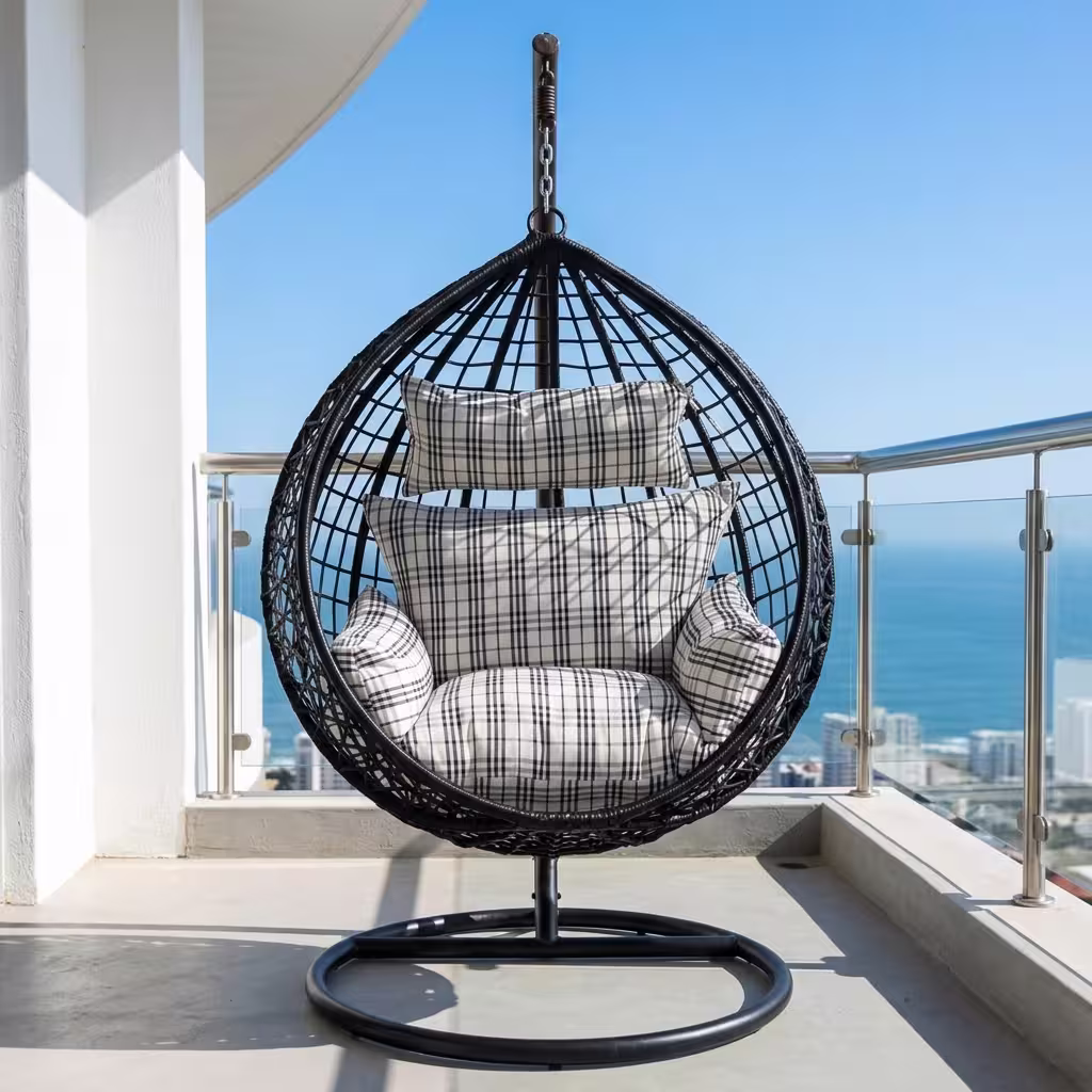 Premium 1-Seater Modern Rattan Hammock Swing featuring a black hand-woven teardrop wicker basket, a heavy-duty dark gray steel hanging stand, and plush black-and-white plaid cushions, placed on a high-rise balcony overlooking a city skyline.