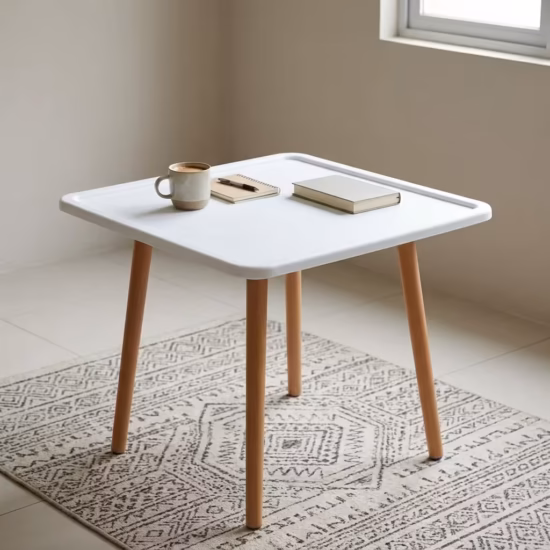 Premium 4-Seater Square Wooden Dining Table in an elegant matte white and natural wood dual-tone finish, showcasing a square tray-style top panel holding a ceramic coffee mug, a leather notebook with a pen, and a closed white hardbound book, supported by four tapered round solid beechwood legs, resting on a textured geometric cream area rug over polished tile flooring.