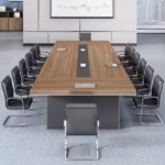 Premium 14-16 Seater Modern Conference Table in a rich walnut woodgrain finish, showcasing a massive rectangular top panel with a center-running black accent band and four silver flip-top power grommets supporting an open laptop, anchored by three heavy-duty matte charcoal gray block leg pillars, resting on a textured blue area rug in a modern office setup with matching wood storage cabinets in the background.