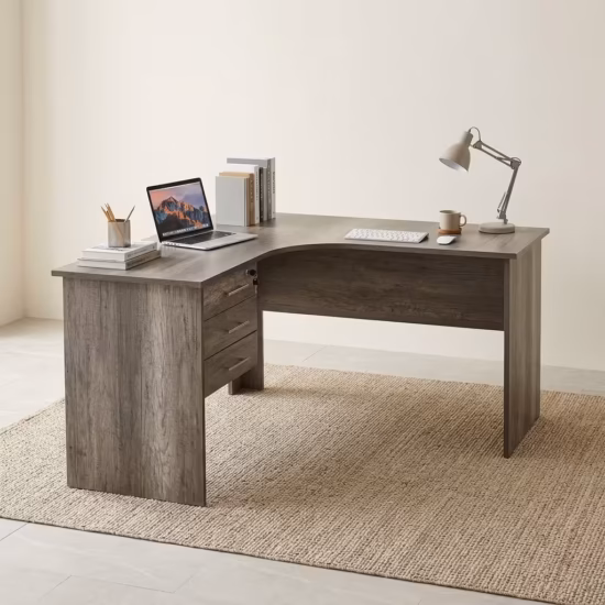 Premium 1.2M Curved Office Desk With Drawers in a warm oak woodgrain finish, showcasing a flat rectangular left-hand curved tabletop supporting a desktop computer monitor, keyboard, mouse, modern desk lamp, and a ceramic mug, with an integrated storage drawer pedestal showing a key lock, resting on a textured light tan area rug.