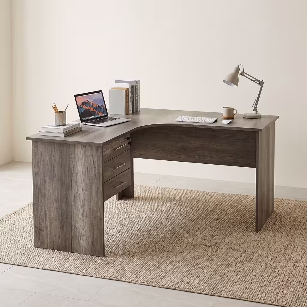 Premium 1.2M Curved Office Desk With Drawers in a warm oak woodgrain finish, showcasing a flat rectangular left-hand curved tabletop supporting a desktop computer monitor, keyboard, mouse, modern desk lamp, and a ceramic mug, with an integrated storage drawer pedestal showing a key lock, resting on a textured light tan area rug.