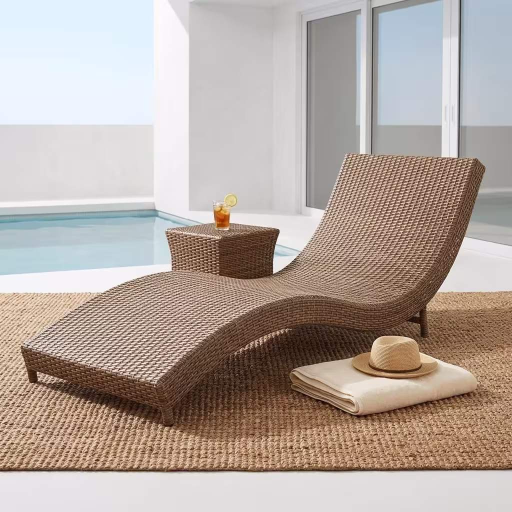 Premium Outdoor Rattan Sun Lounger featuring an ergonomically curved hand-woven natural-tone wicker frame, a plush off-white water-resistant full-length cushion, styled with two blue accent pillows, resting on a textured woven jute rug near a small wooden side table.