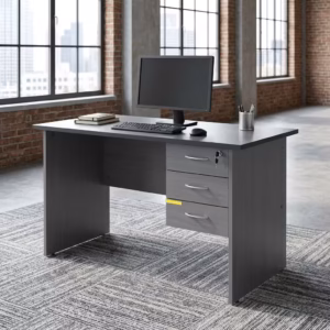 Premium 1.4M Economic Home/Office Study Desk in a dark charcoal grey finish, showcasing a flat rectangular tabletop supporting an open laptop, a cup filled with writing utensils, a mug, and a modern desk lamp on stacked books, with an integrated light grey 3-drawer storage pedestal showing a key lock, resting on a textured grey carpet in front of large windows.