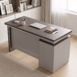 Premium 1200mm Grey Oak Executive Office Desk featuring a rich dark grey oak woodgrain top panel, supported by a front modesty shield in a matte grey finish, an integrated side return storage console block, a desktop supporting an open silver laptop, a white ceramic pen cup, a white desk lamp, and closed journals, resting on a textured woven area rug over light flooring.