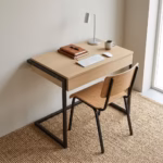 Premium Modern Student Locker Desk in a natural light oak and matte black finish, showcasing a compact rectangular top panel holding a silver keyboard, mouse, leather notebook, ceramic mug, and a sleek silver table lamp, supported by a continuous black metal sled frame base, paired with a matching oak and black metal chair on a thick woven sisal area rug.