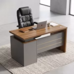 Premium Executive 1.4M Office Desk With Drawers featuring a rich walnut woodgrain top panel, supported by a front modesty shield in a matte grey finish decorated with three horizontal gold accent lines, an integrated left-side return storage console block, a desktop supporting an open silver laptop, a leather notebook with three pens, a ceramic mug, and a closed white book, paired with a high-back black leather executive chair, resting on a textured chevron-patterned woven area rug over a polished concrete floor.