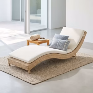 Premium Outdoor Rattan Sun Lounger featuring an ergonomically curved hand-woven natural-tone wicker frame, a plush off-white water-resistant full-length cushion, styled with two blue accent pillows, resting on a textured woven jute rug near a small wooden side table.