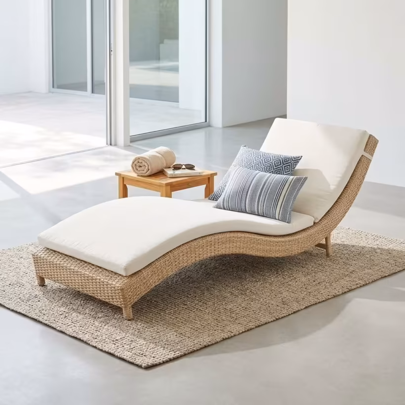 Premium Outdoor Rattan Sun Lounger featuring an ergonomically curved hand-woven natural-tone wicker frame, a plush off-white water-resistant full-length cushion, styled with two blue accent pillows, resting on a textured woven jute rug near a small wooden side table.
