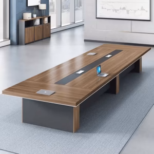 Premium 14-16 Seater Modern Conference Table in a rich walnut woodgrain finish, showcasing a massive rectangular top panel with a center-running black accent band and four silver flip-top power grommets supporting an open laptop, anchored by three heavy-duty matte charcoal gray block leg pillars, resting on a textured blue area rug in a modern office setup with matching wood storage cabinets in the background.