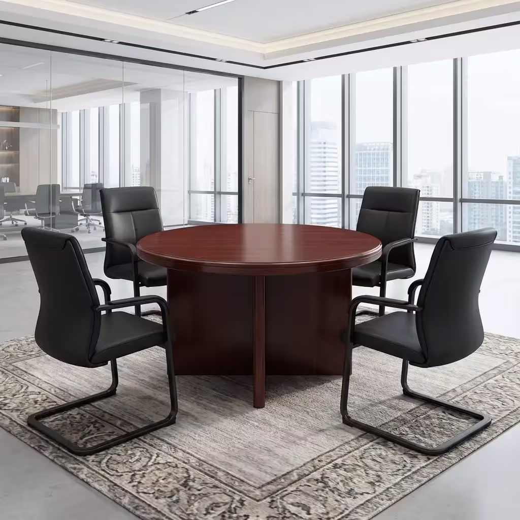 Premium 4-Seater Wooden Meeting Table featuring a thick round top panel in a rich cherry-mahogany woodgrain finish, supported by a heavy-duty centered cross-panel wooden pillar base, surrounded by four black mesh visitor chairs with chrome cantilever sled bases, resting on a smooth grey concrete studio floor next to a massive city-view window.