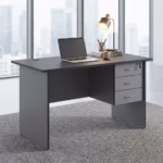 Premium 1.4M Economic Home/Office Study Desk in a dark charcoal grey finish, showcasing a flat rectangular tabletop supporting an open laptop, a cup filled with writing utensils, a mug, and a modern desk lamp on stacked books, with an integrated light grey 3-drawer storage pedestal showing a key lock, resting on a textured grey carpet in front of large windows.