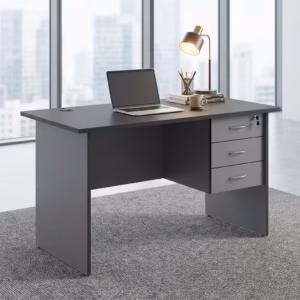 Premium 1.4M Economic Home/Office Study Desk in a dark charcoal grey finish, showcasing a flat rectangular tabletop supporting an open laptop, a cup filled with writing utensils, a mug, and a modern desk lamp on stacked books, with an integrated light grey 3-drawer storage pedestal showing a key lock, resting on a textured grey carpet in front of large windows.