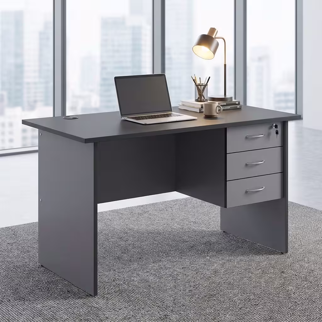 1.4M Economic Home/Office Study Desk Premium 1.4M Economic Home/Office Study Desk in a dark charcoal grey finish, showcasing a flat rectangular tabletop supporting an open laptop, a cup filled with writing utensils, a mug, and a modern desk lamp on stacked books, with an integrated light grey 3-drawer storage pedestal showing a key lock, resting on a textured grey carpet in front of large windows.