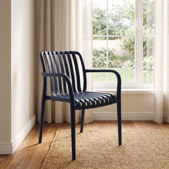 Premium Stackable Plastic Bistro Chair in a deep matte navy blue finish, showcasing a single-piece molded resin frame with an open vertical slatted backrest and seat, sweeping integrated armrests, and clean tapered legs, standing on a textured sisal area rug in a sunlit interior room by a window.