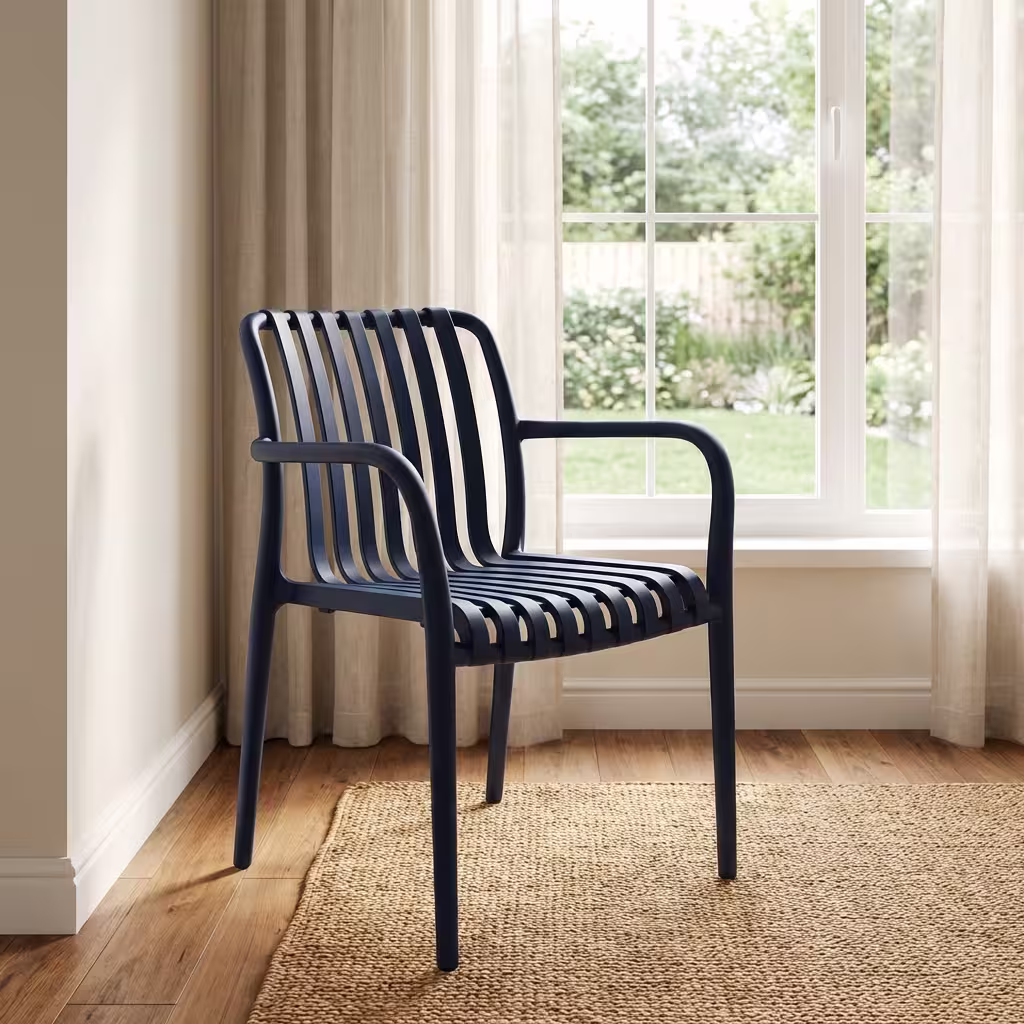 Stackable Plastic Bistro Chair Premium Stackable Plastic Bistro Chair in a deep matte navy blue finish, showcasing a single-piece molded resin frame with an open vertical slatted backrest and seat, sweeping integrated armrests, and clean tapered legs, standing on a textured sisal area rug in a sunlit interior room by a window.