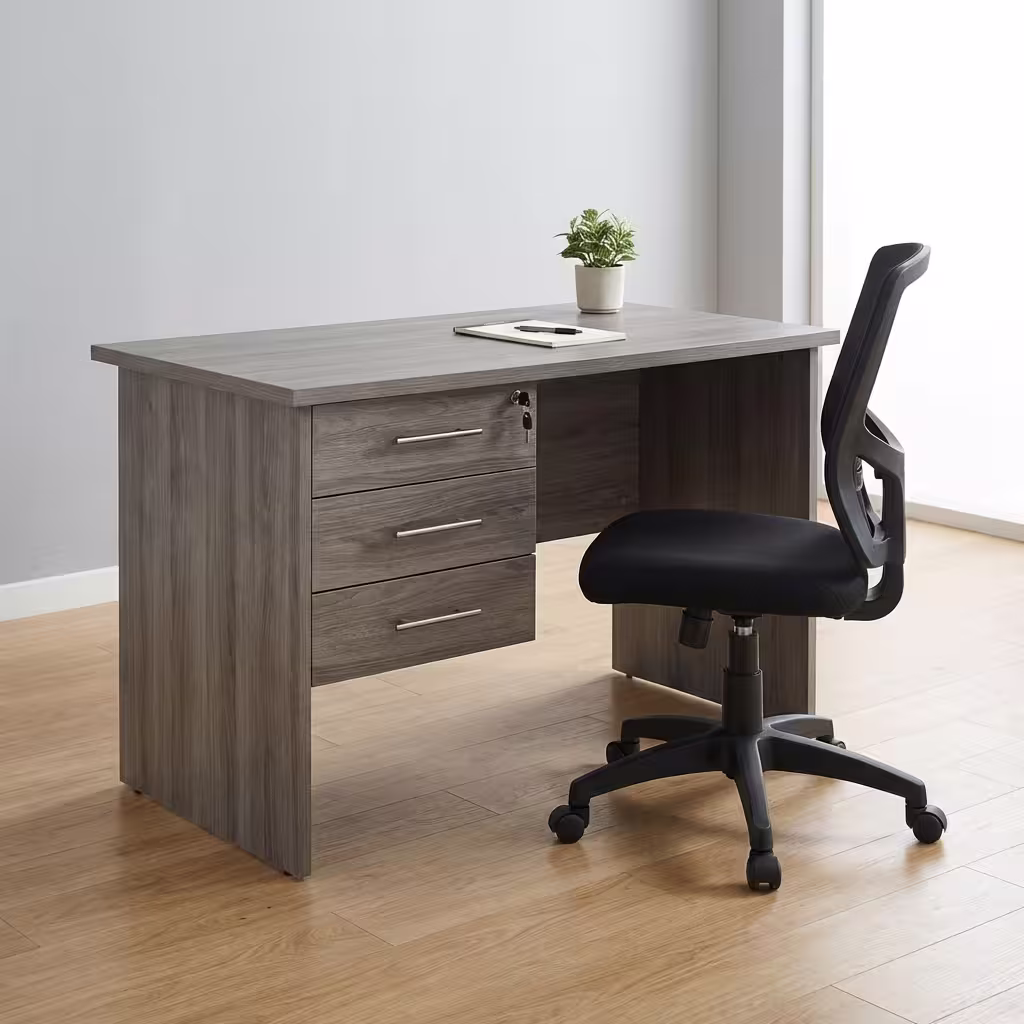 Premium 1.2M Custom Desk With Drawers in a weathered grey woodgrain finish, showcasing a spacious rectangular top panel holding a notebook and a potted plant, an integrated three-drawer left storage pedestal with silver handles and a key inserted in the top lock core, paired with a black mesh office task chair on light wood flooring.