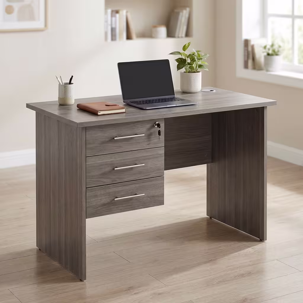 Premium 1.2M Custom Desk With Drawers in a weathered grey woodgrain finish, showcasing a spacious rectangular top panel holding a notebook and a potted plant, an integrated three-drawer left storage pedestal with silver handles and a key inserted in the top lock core, paired with a black mesh office task chair on light wood flooring.
