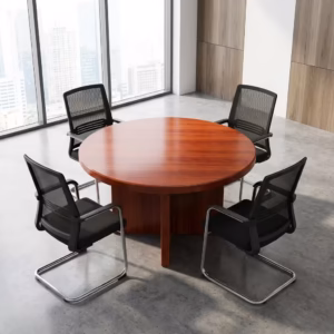 Premium 4-Seater Wooden Meeting Table featuring a thick round top panel in a rich cherry-mahogany woodgrain finish, supported by a heavy-duty centered cross-panel wooden pillar base, surrounded by four black mesh visitor chairs with chrome cantilever sled bases, resting on a smooth grey concrete studio floor next to a massive city-view window.
