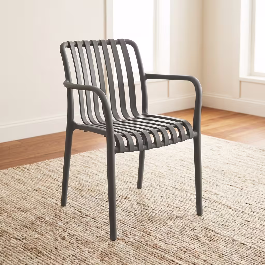 Premium Stackable Plastic Bistro Chair in a deep matte navy blue finish, showcasing a single-piece molded resin frame with an open vertical slatted backrest and seat, sweeping integrated armrests, and clean tapered legs, standing on a textured sisal area rug in a sunlit interior room by a window.