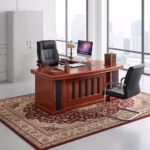 Premium 1.6M Office Desk With Side Extension in a deep cherry-mahogany woodgrain finish, featuring a front modesty panel with vertical black structural columns, an integrated side return storage console supporting an open all-in-one desktop computer, a leather desk blotter mat, a classic stained glass desk lamp, and stacked books, paired with a high-back black leather executive chair and a black leather visitor chair, resting on an ornate patterned traditional area rug.