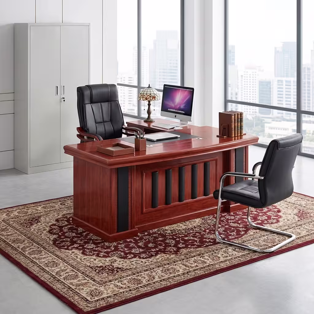 Premium 1.6M Office Desk With Side Extension in a deep cherry-mahogany woodgrain finish, featuring a front modesty panel with vertical black structural columns, an integrated side return storage console supporting an open all-in-one desktop computer, a leather desk blotter mat, a classic stained glass desk lamp, and stacked books, paired with a high-back black leather executive chair and a black leather visitor chair, resting on an ornate patterned traditional area rug.