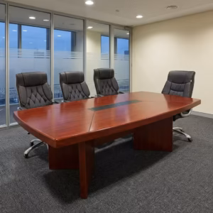 Premium 2.0M Conference Meeting Table in a rich cherry-mahogany woodgrain finish, showcasing an expansive rectangular top panel with tapered ends, a center-running black accent band, and two open flip-top power socket hubs supporting laptops, open notebooks, and coffee cups, anchored by heavy-duty interlocking wooden panel leg bases, surrounded by grey mesh office chairs on a smooth white floor inside a sunlit high-rise boardroom overlooking a city skyline.