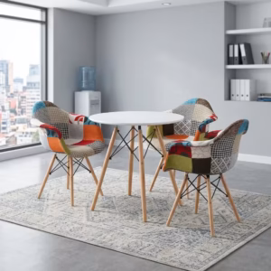Premium Dining Round Table + 3 Chair Set in a contemporary workspace corner, showcasing a sleek round matte white table and three matching bucket armchairs upholstered in a multi-color patchwork fabric with red, teal, orange, and houndstooth patterns, all anchored by tapered light wood legs with black metal cross-wires, resting on a faded blue geometric area rug over polished concrete flooring next to a large window.
