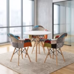 Premium Dining Round Table + 3 Chair Set in a contemporary workspace corner, showcasing a sleek round matte white table and three matching bucket armchairs upholstered in a multi-color patchwork fabric with red, teal, orange, and houndstooth patterns, all anchored by tapered light wood legs with black metal cross-wires, resting on a faded blue geometric area rug over polished concrete flooring next to a large window.