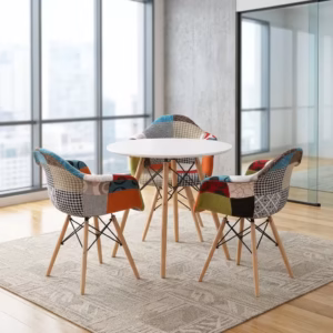 Premium Dining Round Table + 3 Chair Set in a contemporary workspace corner, showcasing a sleek round matte white table and three matching bucket armchairs upholstered in a multi-color patchwork fabric with red, teal, orange, and houndstooth patterns, all anchored by tapered light wood legs with black metal cross-wires, resting on a faded blue geometric area rug over polished concrete flooring next to a large window.
