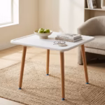 Premium 4-Seater Square Wooden Dining Table in an elegant matte white and natural wood dual-tone finish, showcasing a square tray-style top panel holding a ceramic coffee mug, a leather notebook with a pen, and a closed white hardbound book, supported by four tapered round solid beechwood legs, resting on a textured geometric cream area rug over polished tile flooring.