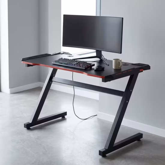 Premium Gaming Desk With Led Light in a sleek matte black carbon fiber texture, showcasing a stable Z-shaped black metal frame, an expansive desktop holding a widescreen curved monitor, mechanical gaming keyboard, mouse, and a beige mug, standing on a polished concrete floor.