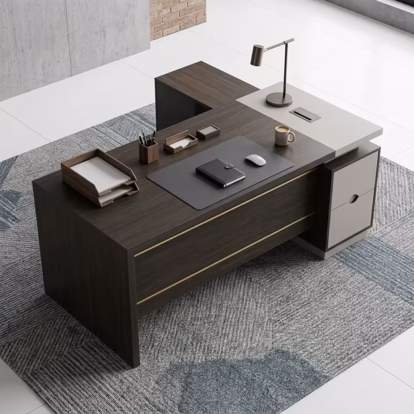 Premium 1.6M L-Shaped Executive Office Desk in a deep charcoal-brown woodgrain finish, featuring a front modesty panel with two horizontal gold accent lines, an integrated stone-grey side return shelf console with an adjustable desk lamp, a flush charcoal leather desk blotter supporting a closed journal and a wireless mouse, and an under-desk storage drawer cabinet, resting on a textured geometric blue-and-grey pattern area rug.