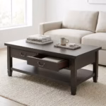 Premium Retro Wooden Coffee Table With Storage in a deep charcoal-brown woodgrain finish, showcasing a rectangular top panel holding stacked books and a ceramic mug on a marble tray, a front section with one closed drawer and one partially open drawer displaying dark metal antique handles, an open lower storage shelf, resting on a thick textured cream area rug in front of a beige fabric sofa.