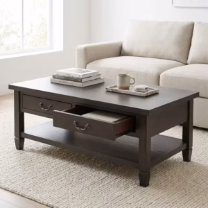 Premium Retro Wooden Coffee Table With Storage in a deep charcoal-brown woodgrain finish, showcasing a rectangular top panel holding stacked books and a ceramic mug on a marble tray, a front section with one closed drawer and one partially open drawer displaying dark metal antique handles, an open lower storage shelf, resting on a thick textured cream area rug in front of a beige fabric sofa.